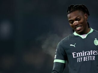Footballeur souriant en maillot sombre, fond flou.