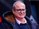Homme aux lunettes et manteau marron, souriant.