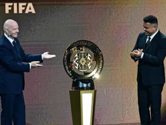 Deux hommes dévoilent un trophée FIFA sur scène.