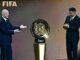 Deux hommes dévoilent un trophée FIFA sur scène.