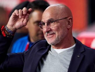Homme souriant avec lunettes et veste, saluant.