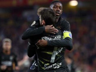 Deux footballeurs en maillot noir s’embrassent sur le terrain
